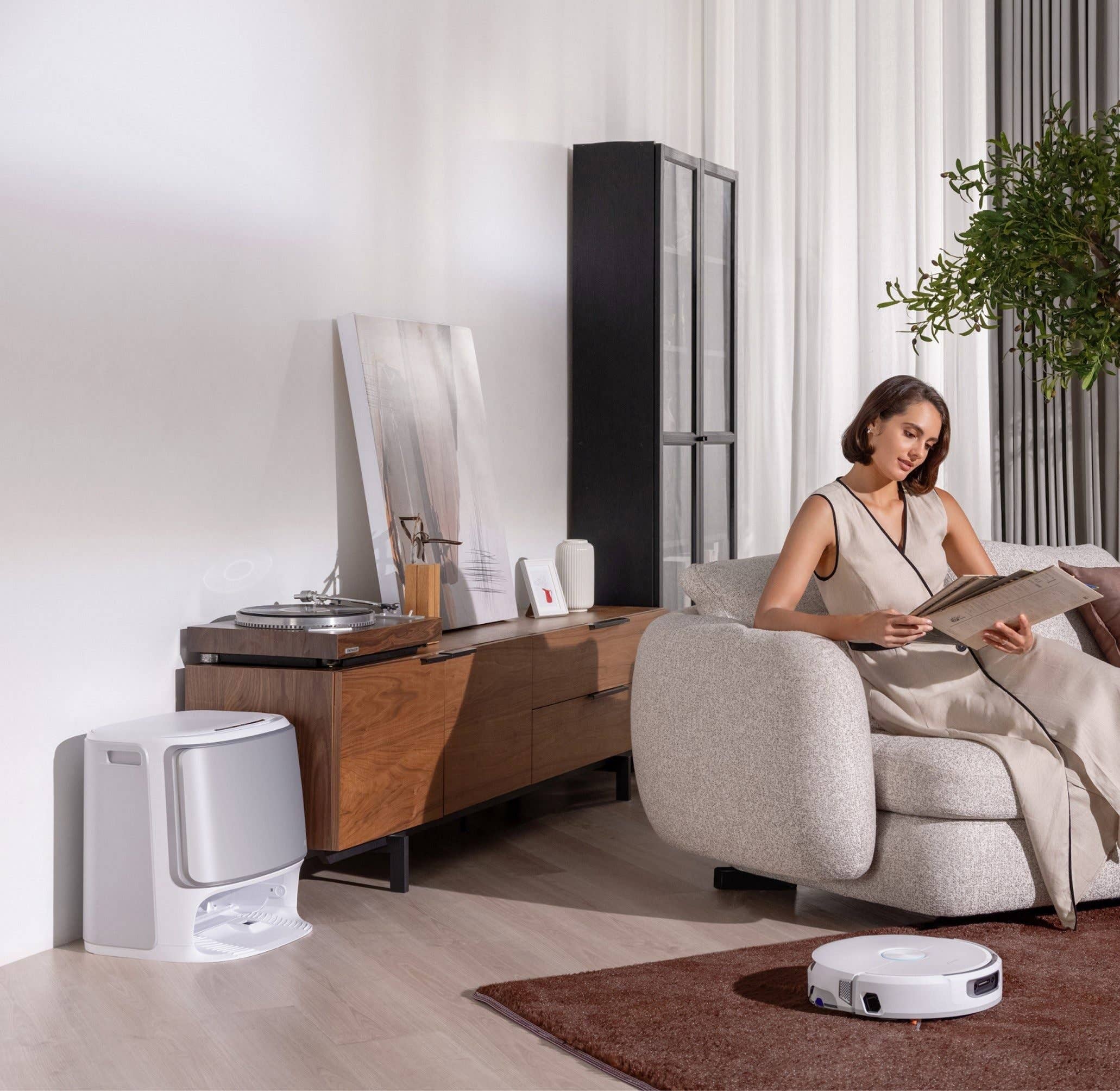 A woman reading a magazine while sitting on a sofa, with a robotic vacuum cleaner on the floor.