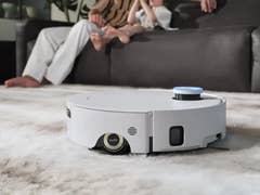 A Dreame Aqua10 Roller Complete robot vacuum cleaning a patterned rug in a living room with people in the background.