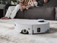 A Dreame Aqua10 Roller Complete robot vacuum cleaning a patterned rug in a living room with people in the background.