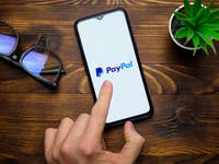 A hand pointing at a smartphone displaying the PayPal logo, with glasses and a small plant nearby.