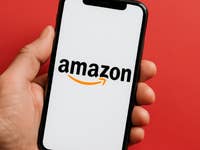 A hand holding a smartphone displaying the Amazon logo on a white screen against a red background.