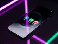 A smartphone displaying three messaging app icons: Telegram, WhatsApp, and Signal on a reflective surface with neon lights.