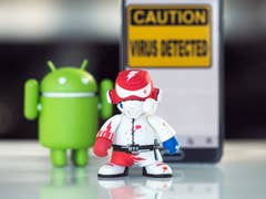 An Android figure and a malware character stand in front of a sign that says "Caution: Virus Detected."