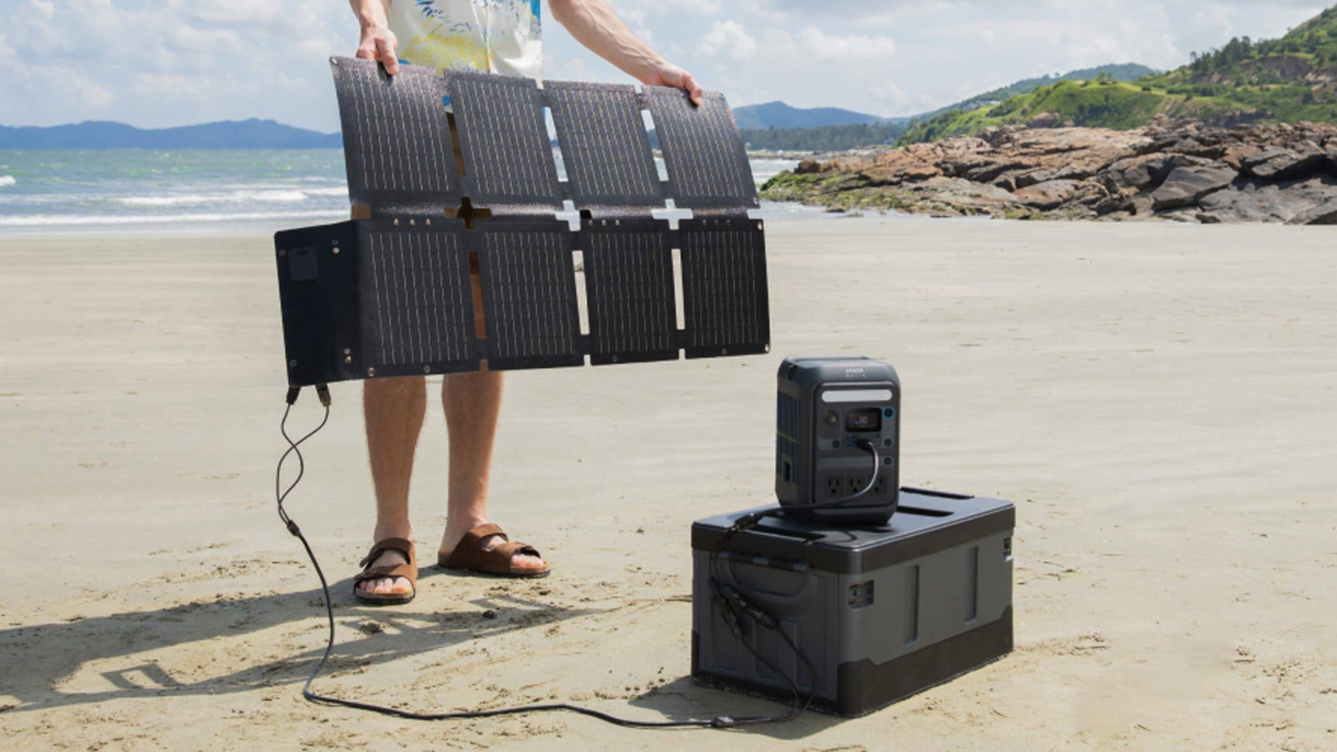 A person holds a folding solar panel over a portable power station on the beach.