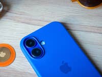 A blue smartphone beside an orange Apple AirTag and yellow earbuds on a wooden surface.