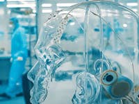 A transparent human head model with wires and sensors in a lab, with a person in protective gear in the background.