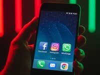 A hand holding a smartphone displaying Facebook, Instagram, WhatsApp, and other apps on the screen.