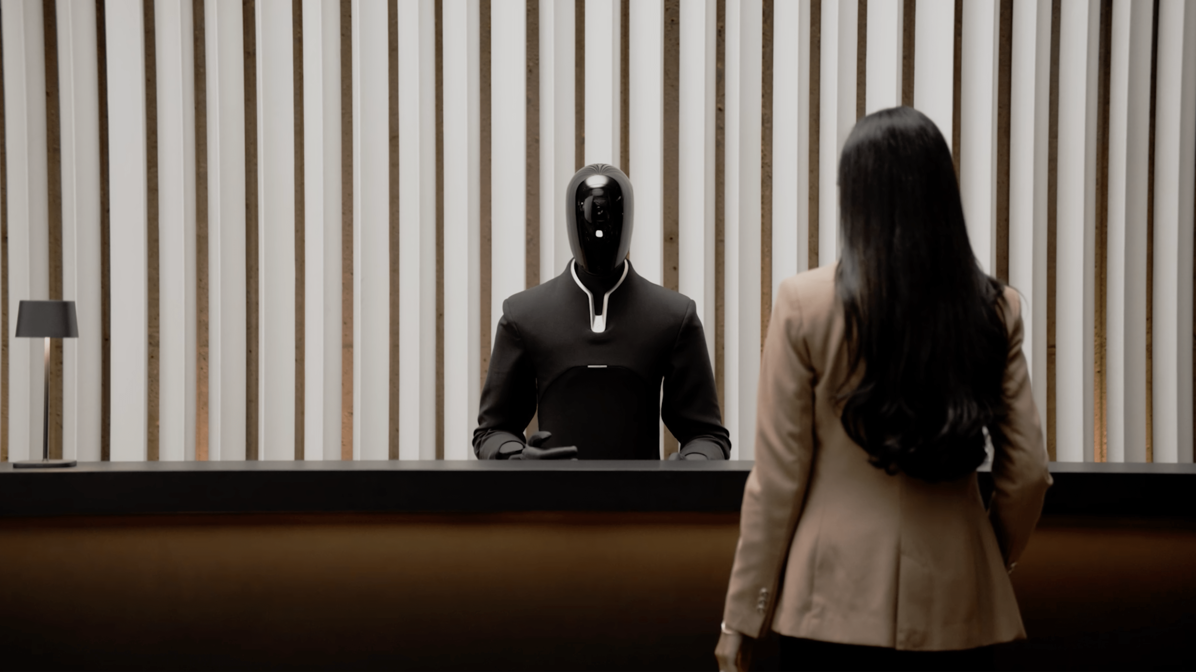A humanoid robot at a reception desk interacting with a woman in a suit.