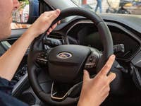 A person holds the steering wheel of a Ford car, with a dashboard and side mirror visible.