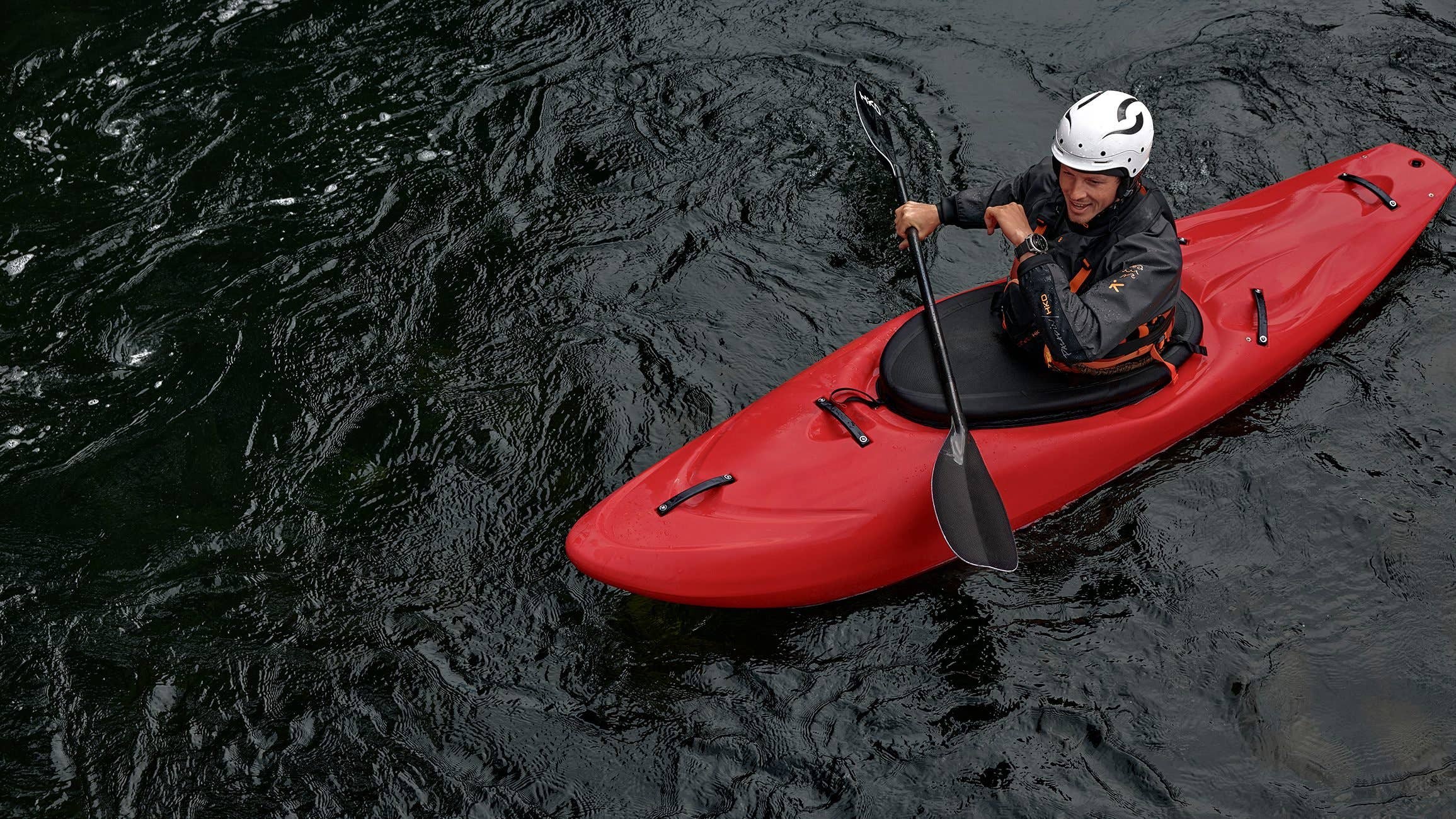 A person paddling a red kayak on dark water, wearing a helmet and a wetsuit.