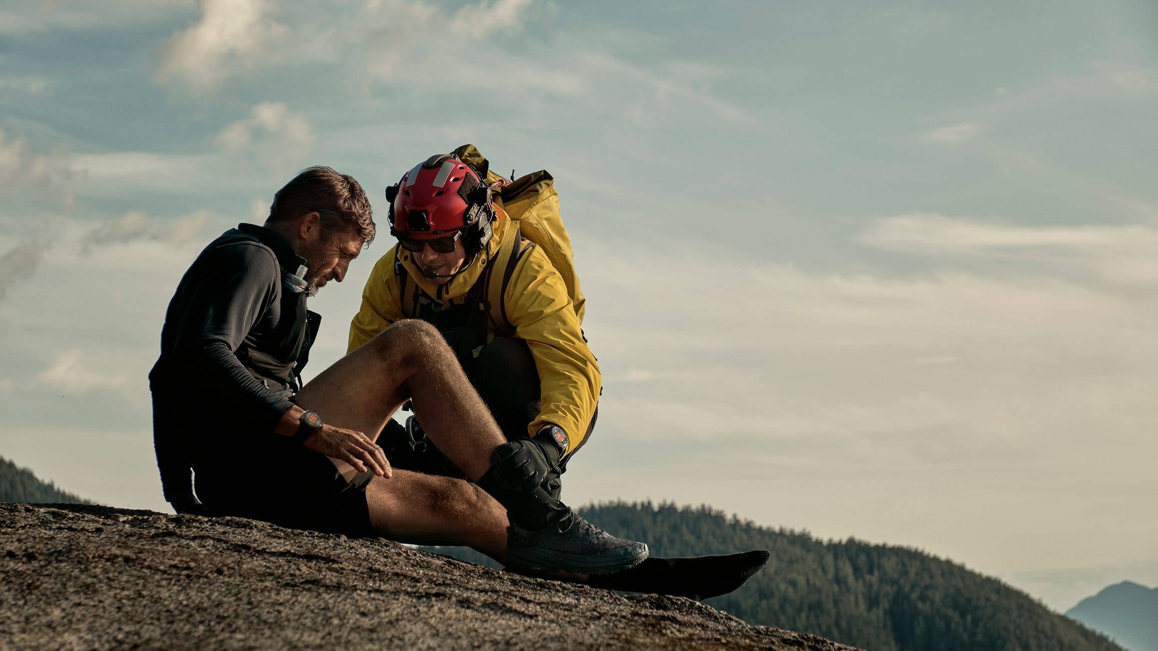 Two climbers assist each other on a rocky surface with mountains in the background.