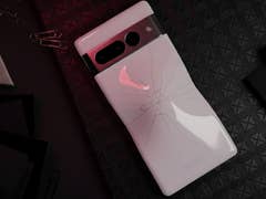 A Google Pixel 7 Pro smartphone with a cracked back, resting on a textured black surface with paper clips and stationery.