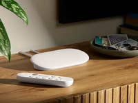 A Google TV 4K streaming device and remote on a wooden table next to a plant and a decorative plate.