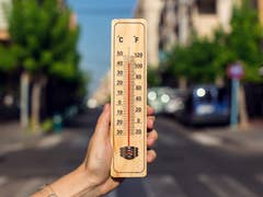 A hand holding a wooden thermometer showing high temperatures in Celsius and Fahrenheit on a sunny street.