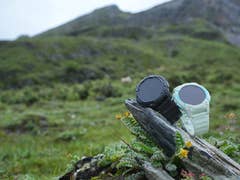 Two Kospet S2 smartwatches, one black and one white, displayed on a rock in a mountainous, grassy landscape.