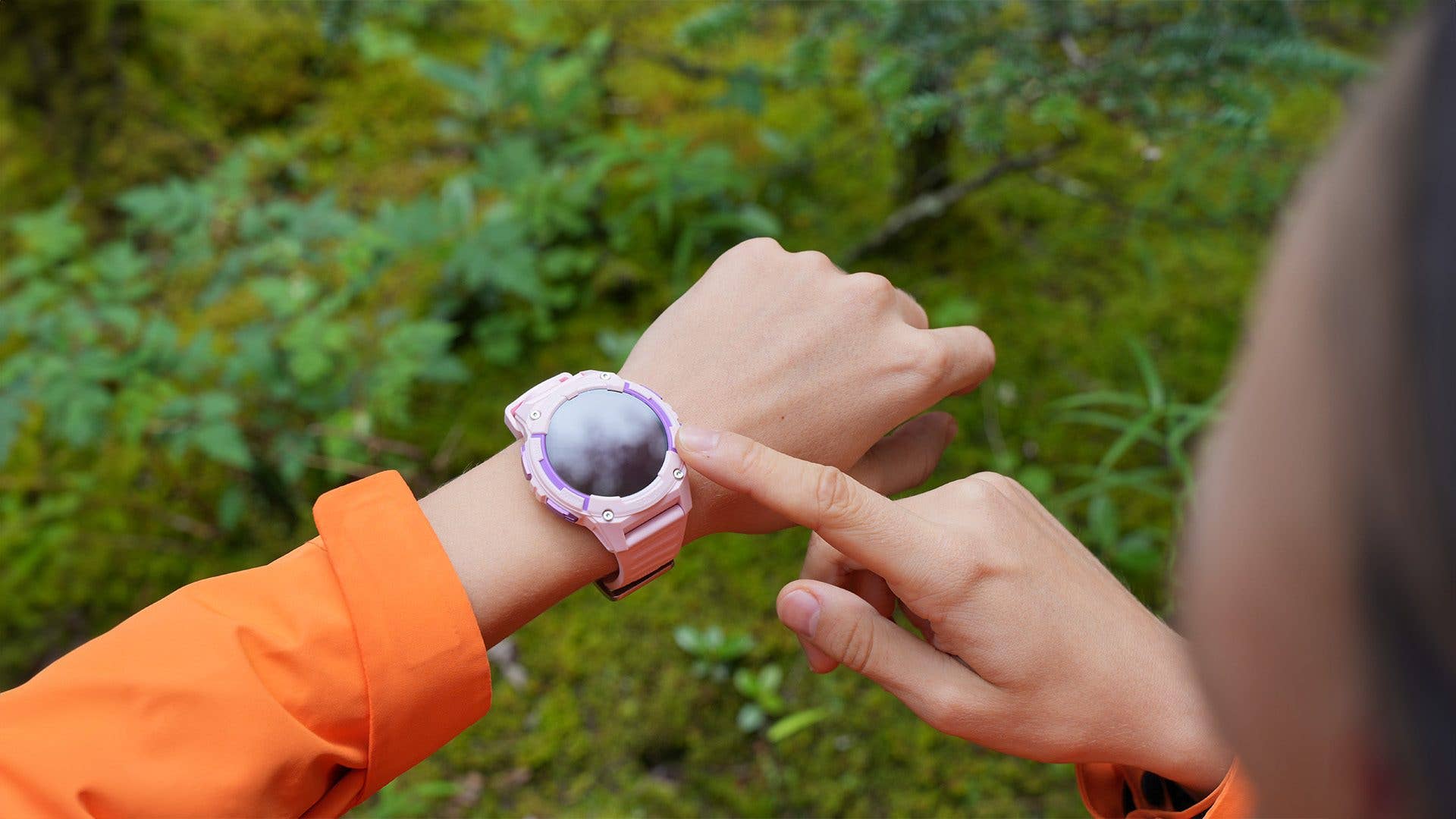 A person wearing an orange jacket touches a Kospet S2 smartwatch on their wrist in a green outdoor setting.