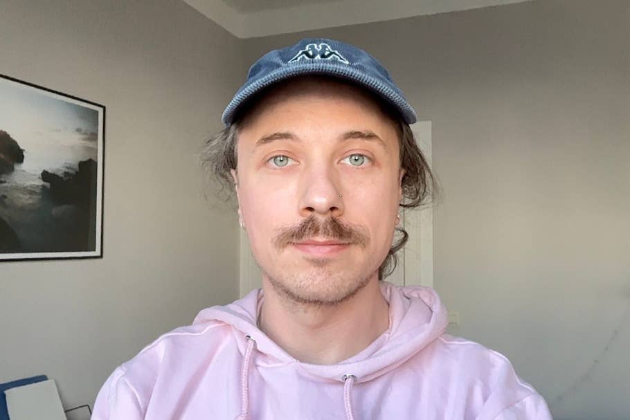 A person with a moustache wearing a pink hoodie and a blue cap against a neutral background.