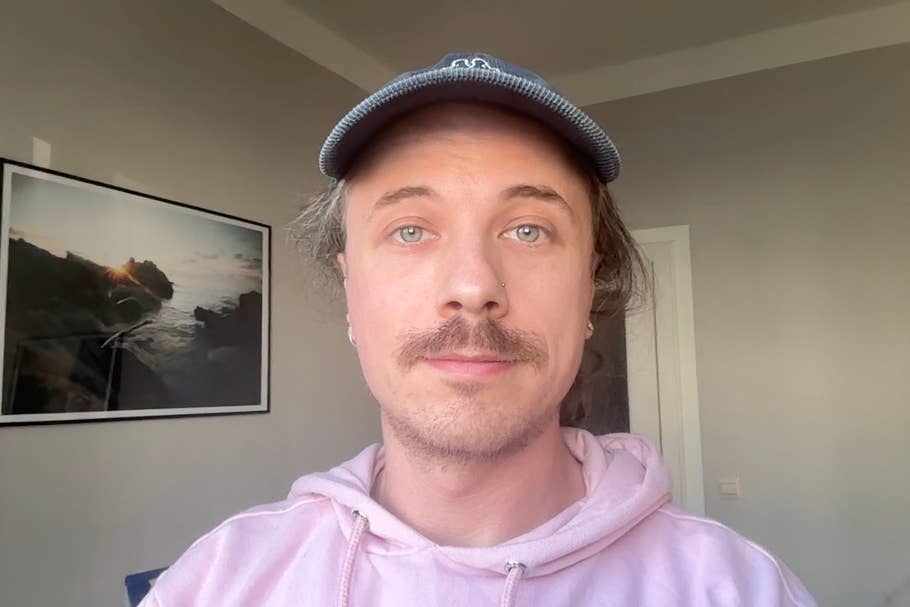 A person with a mustache and a cap wearing a pink hoodie poses in a well-lit room.