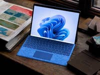 A Microsoft Surface Pro 11 laptop on a wooden table next to fabric swatches and a color sample book.
