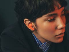 A person with short hair wearing orange earbuds, looking thoughtfully to the side.