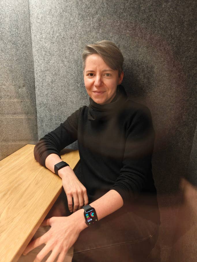 A person sitting at a wooden table, wearing a smartwatch and looking at the camera in a gray-walled room.