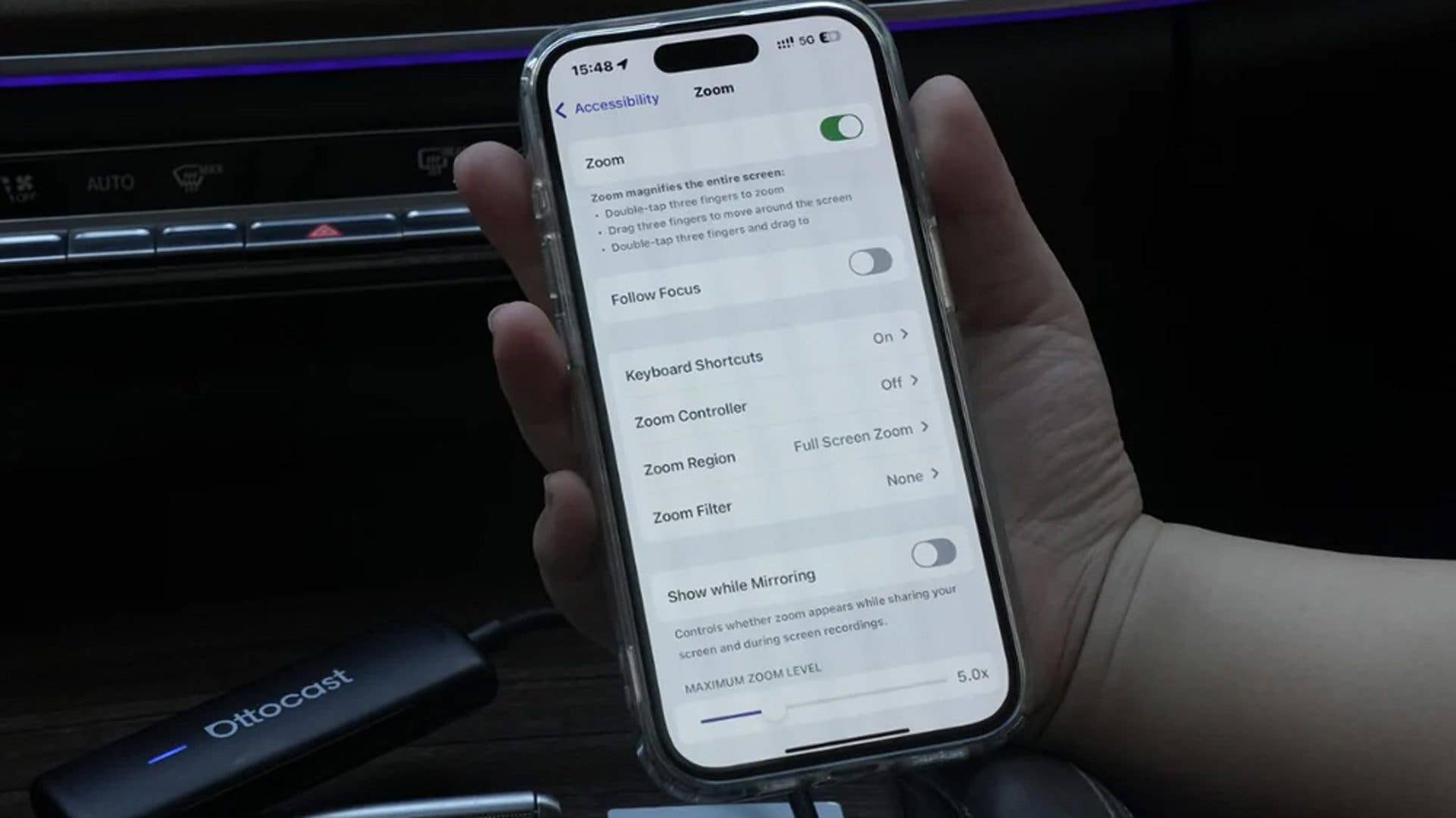 A hand holding a phone displaying Zoom settings under Accessibility, with an Ottocast device nearby.