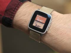 A wrist wearing a Pebble Time smartwatch displaying time and location.