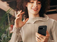 A person with curly hair holds a Google Pixel 9a smartphone and smiles while interacting with it.