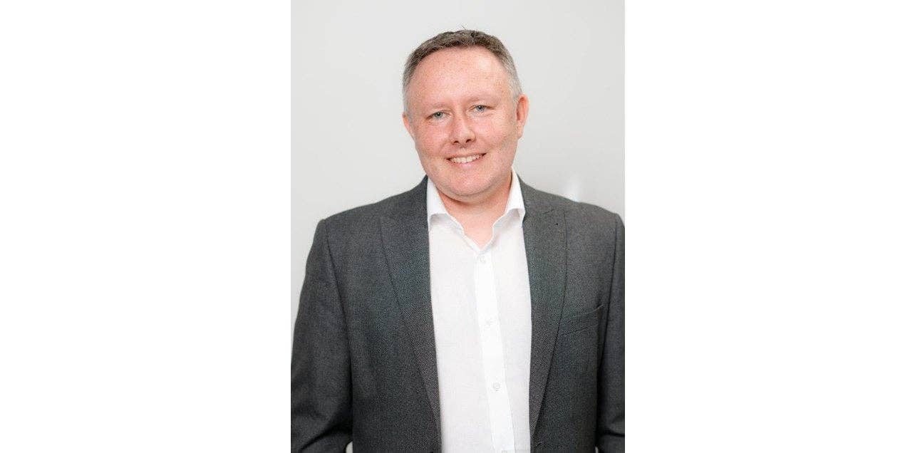 A smiling man in a gray suit and white shirt, standing against a light background. A smiling man in a gray suit and white shirt, standing against a light background.