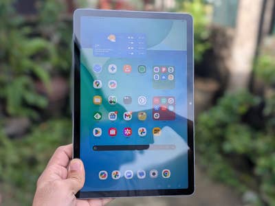 A person holding a Samsung Galaxy Tab S10 FE displaying various app icons on its screen.
