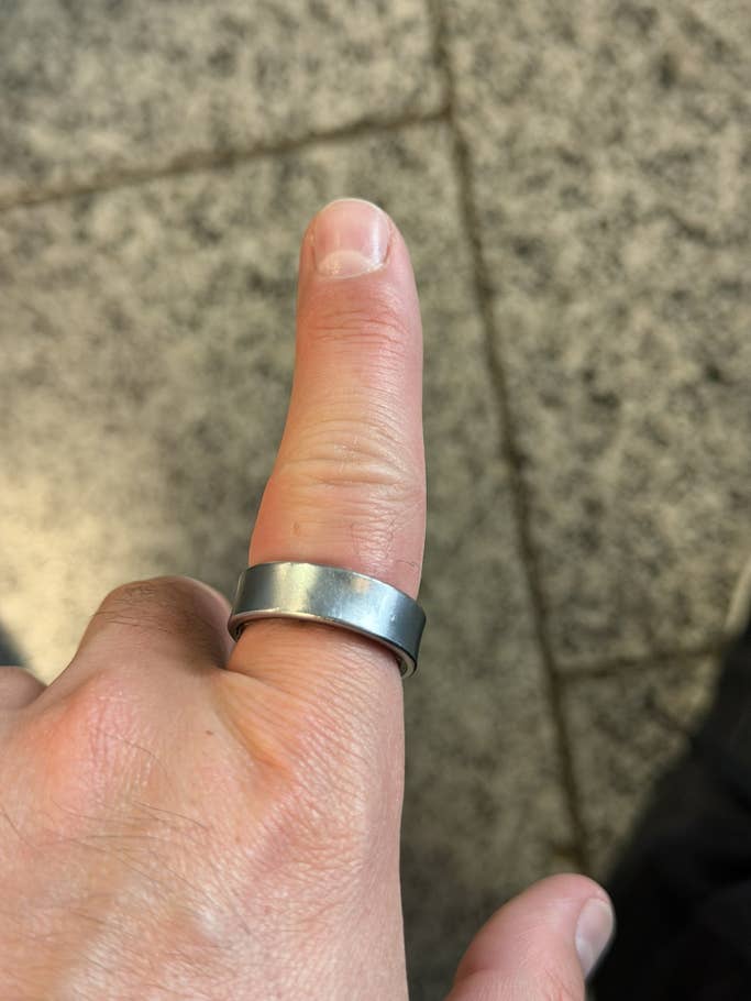 A hand with a metal Samsung Galaxy ring on the thumb, against a textured surface.