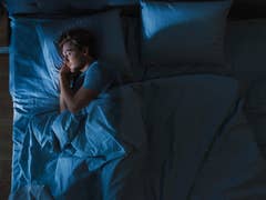 A person sleeping in a bed with blue sheets and pillows in a dimly lit room.