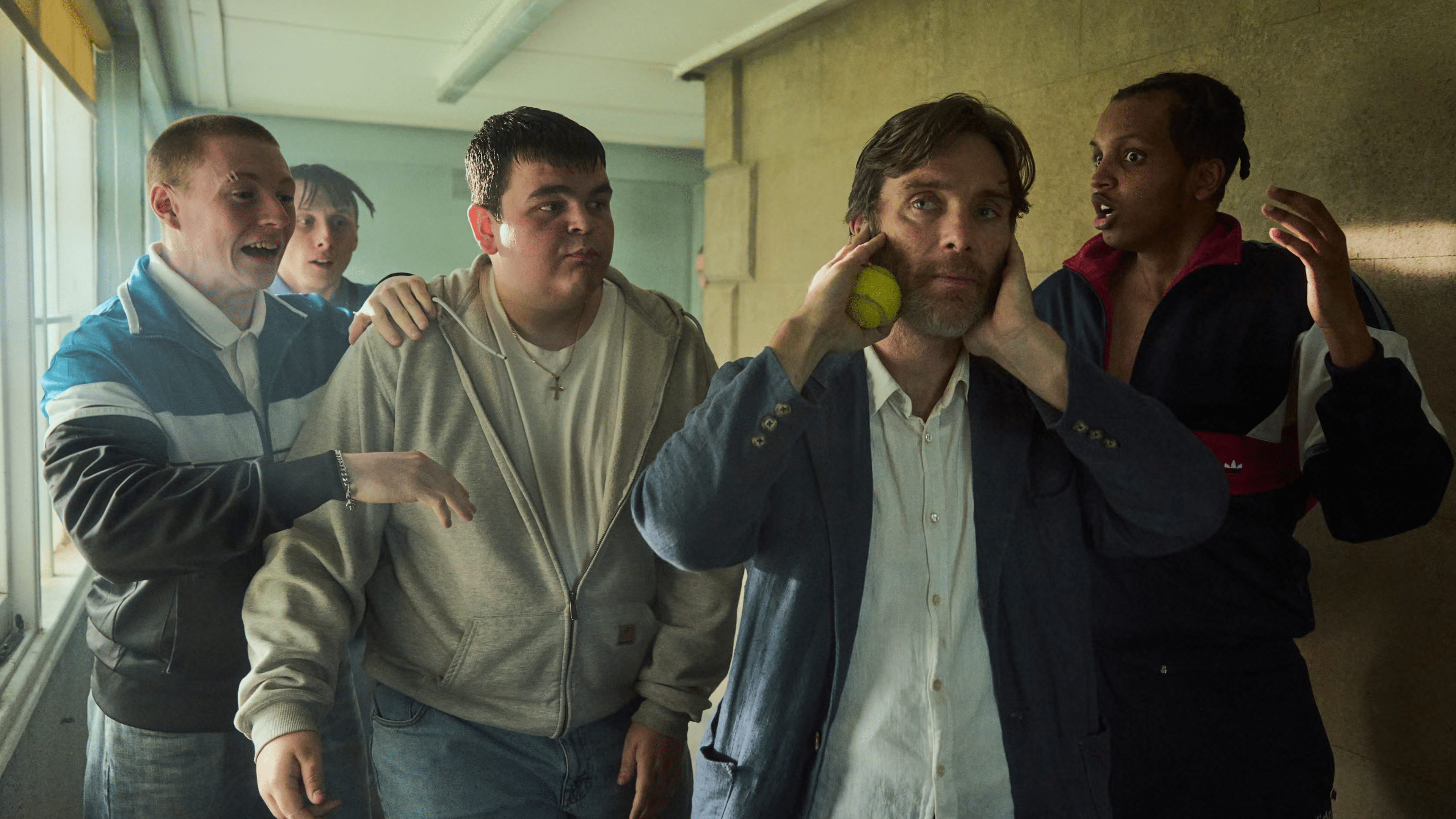 A group of four young men surrounds a concerned man holding his head, with one throwing a tennis ball.