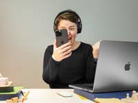 Person with headphones excitedly holding a smartphone in front of a laptop.