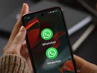 A hand holding a smartphone displaying the WhatsApp app icon twice on the screen.