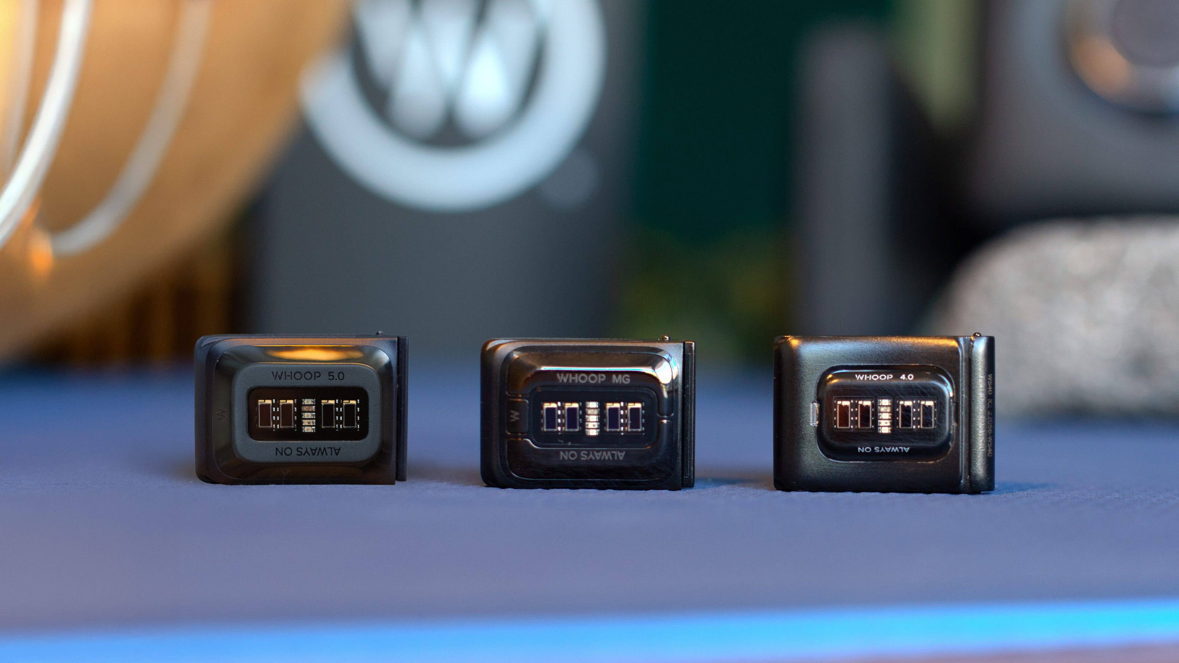 Three WHOOP devices: 5.0, MG, and 4.0, displayed on a blue surface.