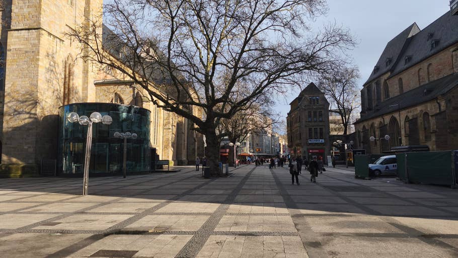 A wide street with pedestrians, flanked by buildings and a large tree, under a clear sky.