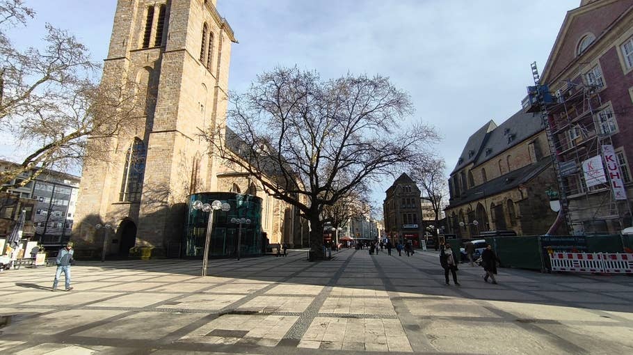 A sunny town square with a historic church, trees and passers-by.