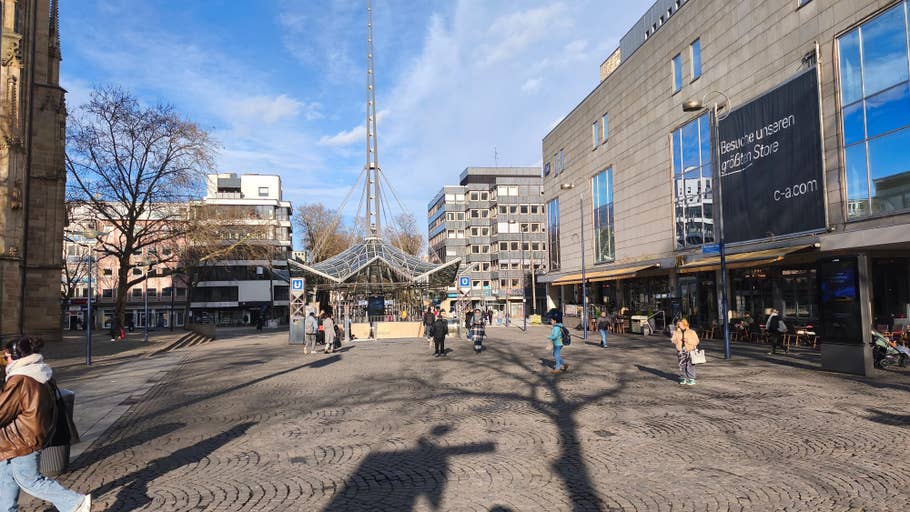A busy town square with passers-by, shops and a glass structure under a blue sky.