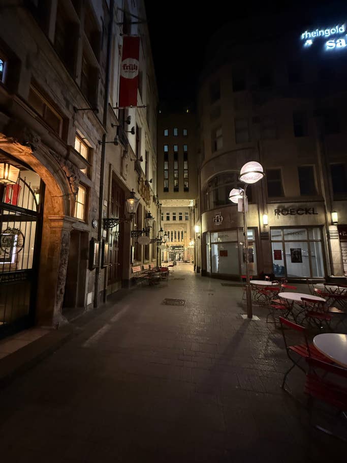 A narrow cobblestone street at night with illuminated buildings and empty red chairs.