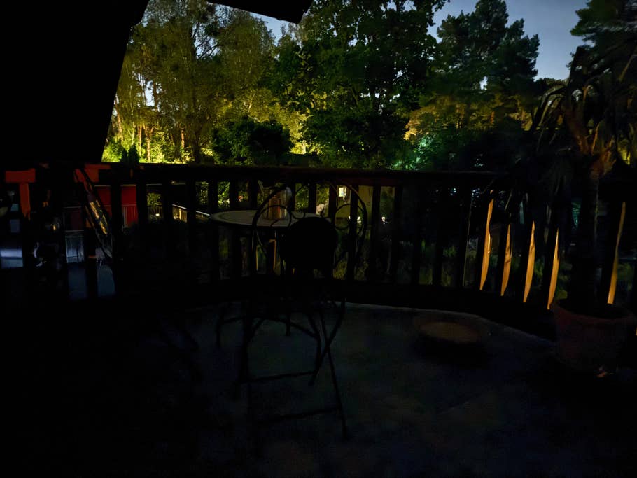 A dimly lit balcony with a table and a chair overlooking a garden at night.