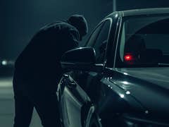 A figure in a hoodie leans over a car window at night, suggesting potential theft.