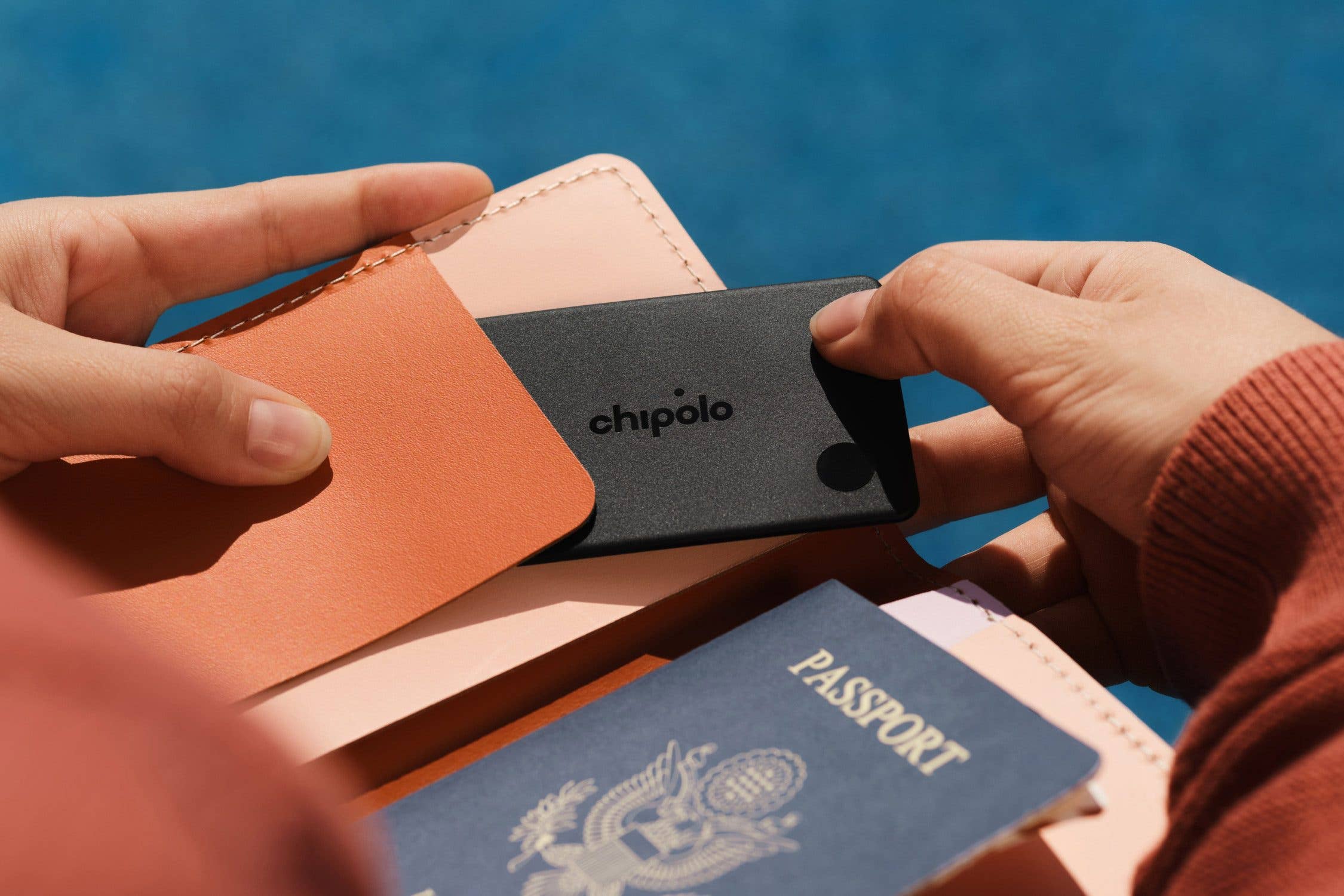 A person holds a Chipolo card item tracker in their hand, next to a passport and wallet.