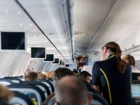 Interior of an airplane cabin with rows of seats and overhead screens, a flight attendant serving passengers.