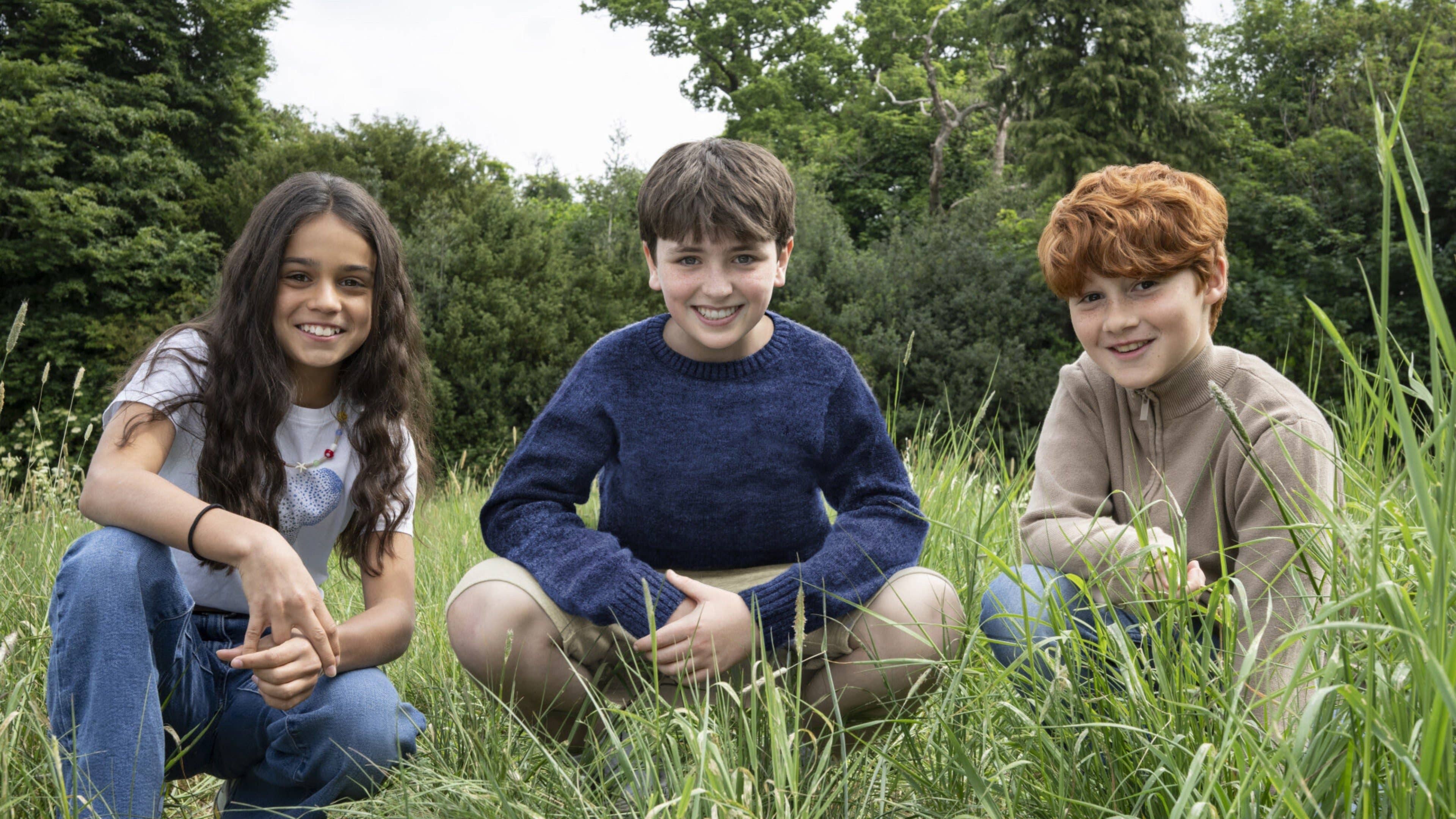 Three children smiling while sitting in grass, with trees in the background.