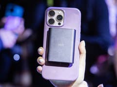 A person holding a smartphone with a black accessory attached to the back in a crowded setting.