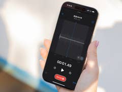 A hand holding an Apple iPhone 16 Pro displaying a voice recording screen with waveform and timer.