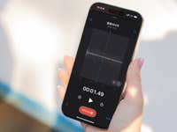 A hand holding an Apple iPhone 16 Pro displaying a voice recording screen with waveform and timer.