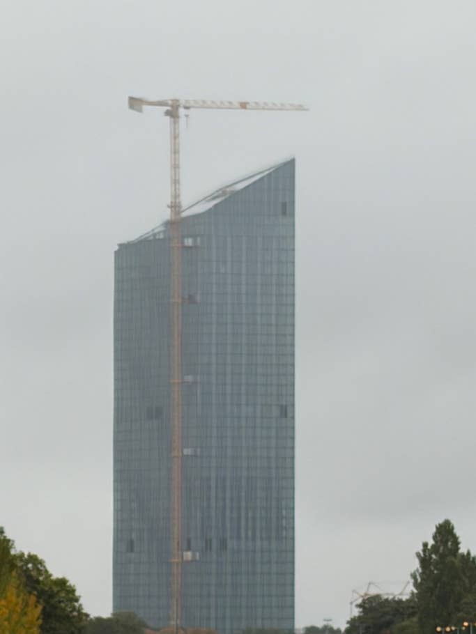 A tall glass tower under construction with a crane on top on a cloudy day.