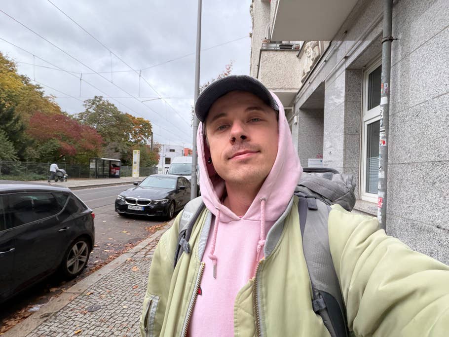 A person stands on a street with a backpack, wearing a pink hoodie and a green jacket.